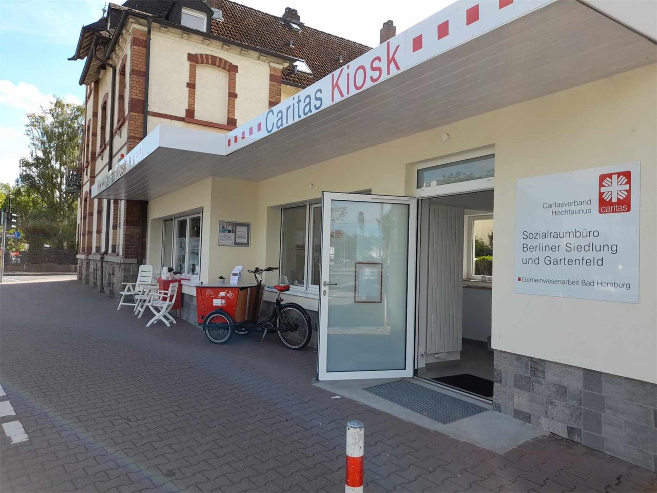 Das Bild zeigt die Außenansicht des "Caritas Kiosk" in Bad Homburg. Vor dem Eingang stehen ein Fahrrad und einige Stühle, was eine einladende und gemeinschaftsorientierte Atmosphäre vermittelt.