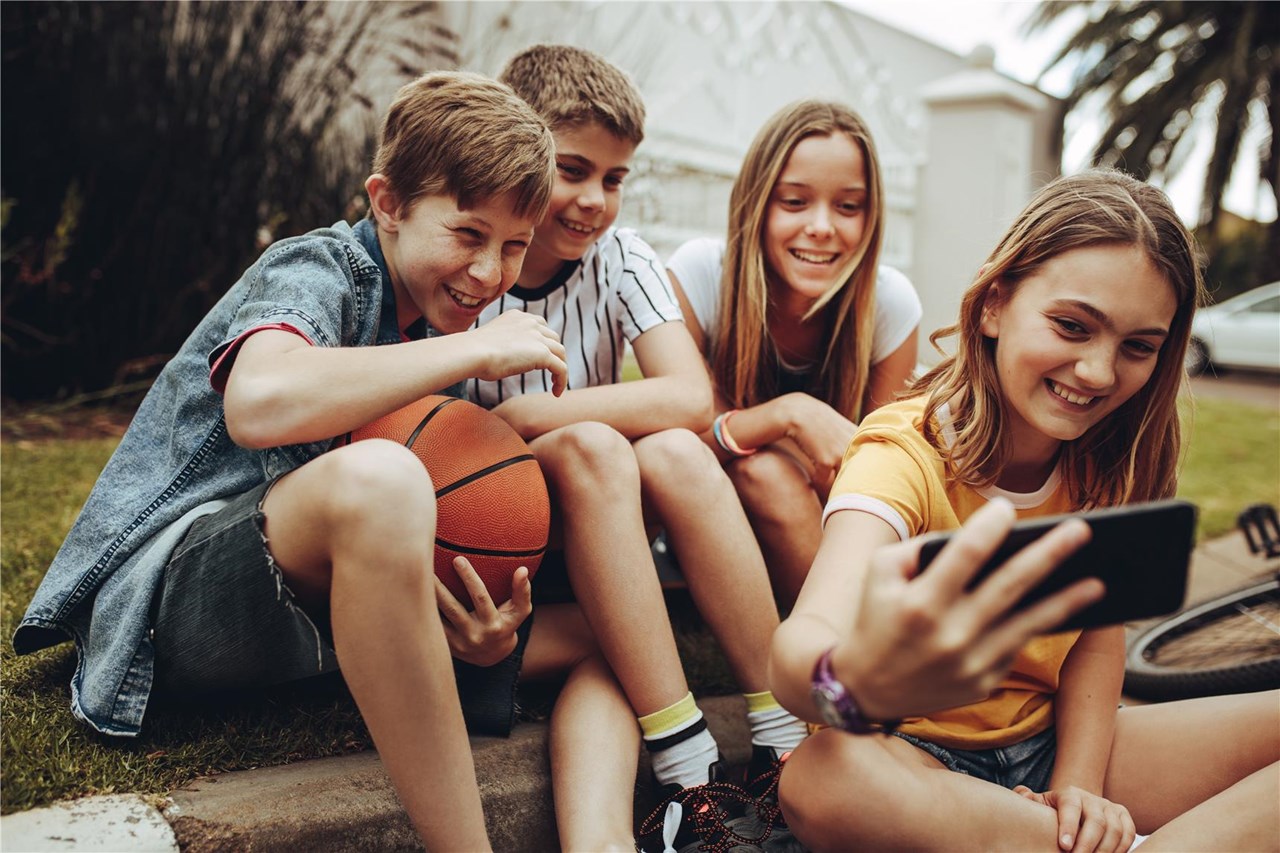 Eine Gruppe von vier Kindern sitzt auf einem Bordstein, lächelt und macht ein Selfie mit einem Smartphone. Einer der Jungen hält einen Basketball und im Hintergrund ist ein Fahrrad zu sehen.